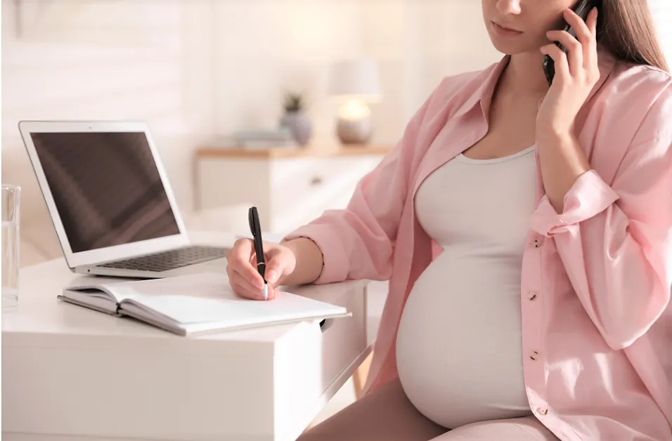 A pregnant woman sits at a desk, talking on the phone—possibly with a Pregnancy Discrimination Lawyer—writing in a notebook, with a laptop and a glass of water nearby.