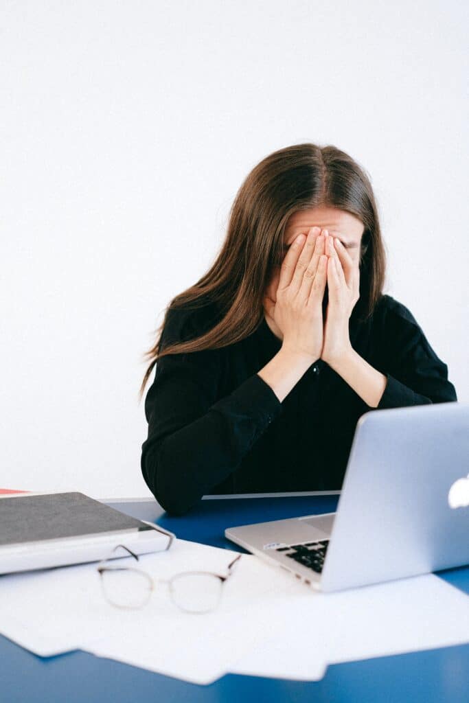 A woman covering her face with her hands in front of a laptop.