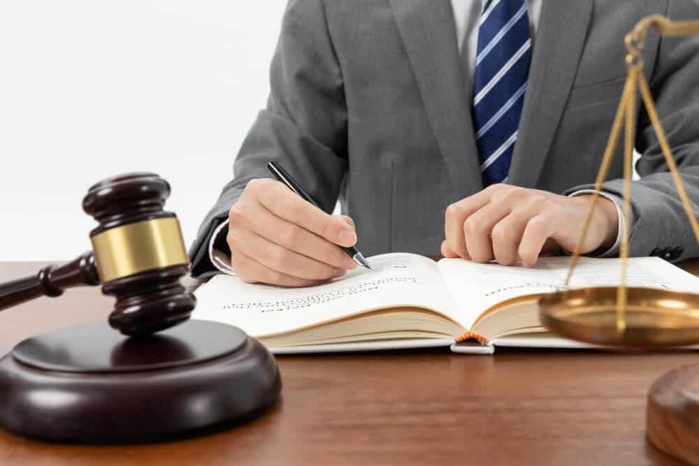 A person in a suit writes in a book at a desk with a gavel and scales of justice.