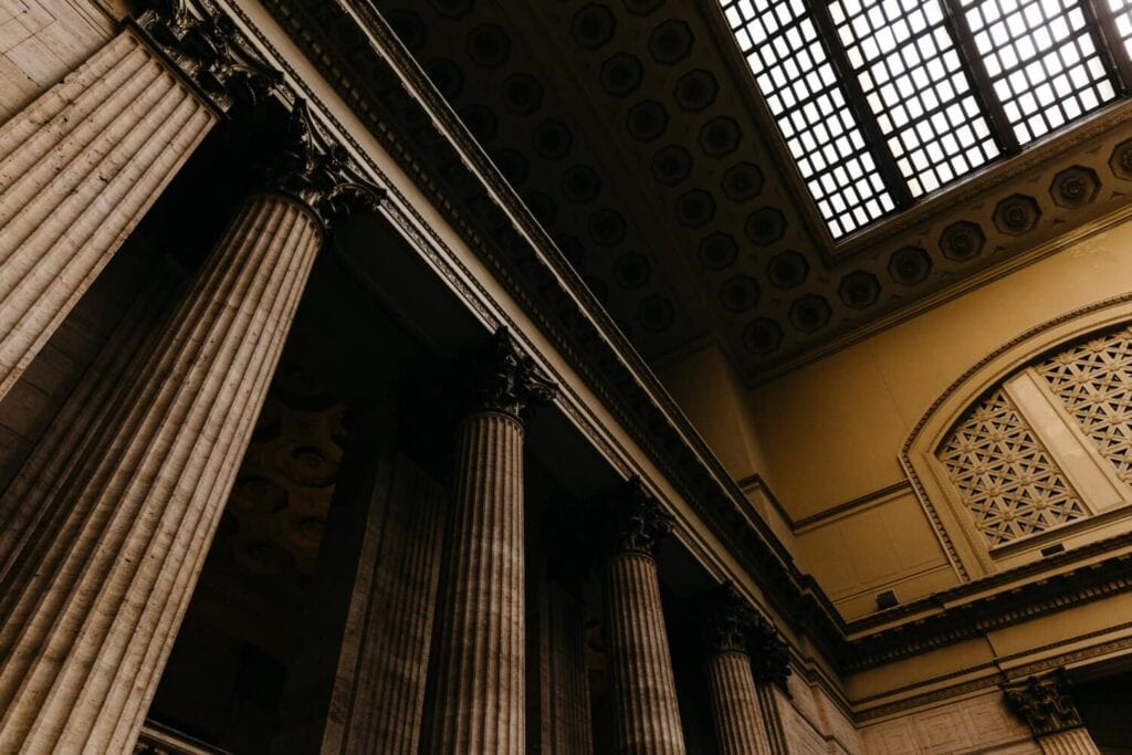 Interior view of a grand building with tall, ornate columns and a large, decorative ceiling featuring a grid pattern skylight.
