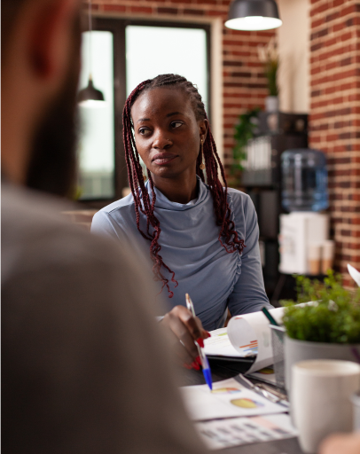 Woman with braided hair sits at a desk holding a pen, looking towards a colleague in an office with exposed brick walls, appearing concerned about possible harassment in what may be a hostile work environment.