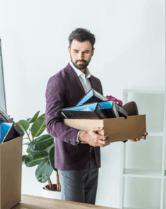 A man in a suit holds a cardboard box filled with office supplies, standing in an office setting near a plant and empty shelves—an image that suggests the impact of harassment or a hostile work environment.