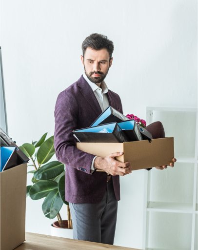 A man in a suit holds a cardboard box filled with office supplies, standing in an office setting near a plant and empty shelves—an image that suggests the impact of harassment or a hostile work environment.