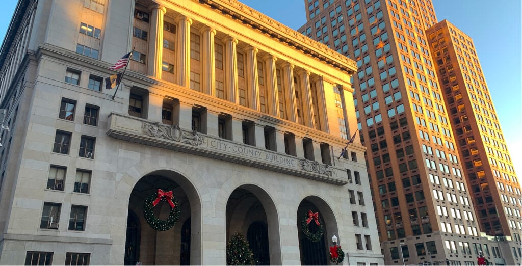 Exterior of a stone city-county building with large arches, columns, American flag, and holiday wreaths—perfect inspiration for your New Homepage—set against a tall, sunlit office building.