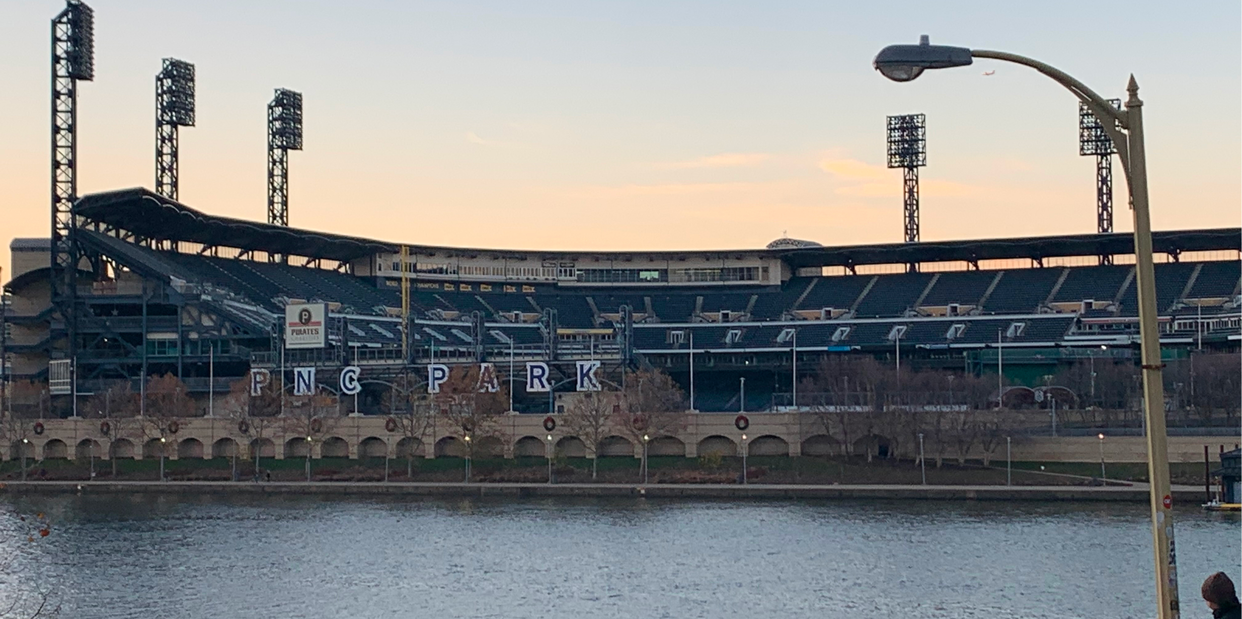 Homepage view of PNC Park, a baseball stadium with empty seats, towering lights, and a river in the foreground under a partly cloudy sky.