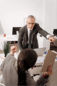 An older man in a suit points and appears to scold a seated woman holding a clipboard, who raises her fist defensively—an office scene suggesting possible workplace discrimination.