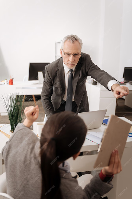 An older man in a suit points and appears to scold a seated woman holding a clipboard, who raises her fist defensively—an office scene suggesting possible workplace discrimination.