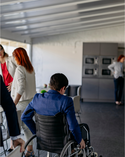 A person in a wheelchair is seated at a table in a modern office space, with three others standing and talking in the background, highlighting the importance of disability rights and inclusion in the workplace.