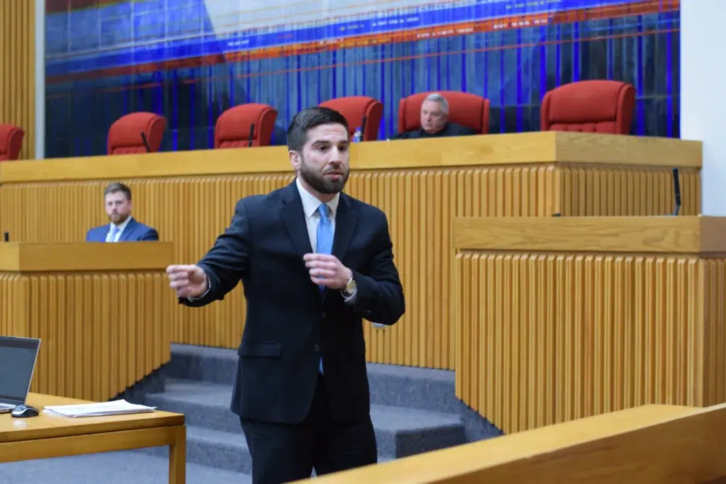 A man in a suit speaks while gesturing in a courtroom, representing the Lacy Employment Law Firm. Seated individuals are visible behind wooden panels in the background.