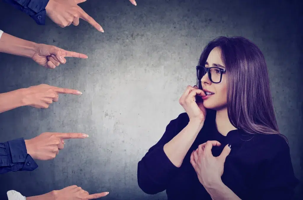 A woman stands nervously biting her nails as multiple hands point at her against a gray background, capturing the anxiety someone might feel before contacting a Retaliation Lawyer.