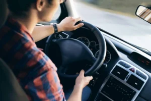 Person driving a car, hands on the steering wheel, dashboard and speedometer visible, wearing a plaid shirt—highlighting the everyday reality for many who face car insurance discrimination or struggle with high rates due to low income.
