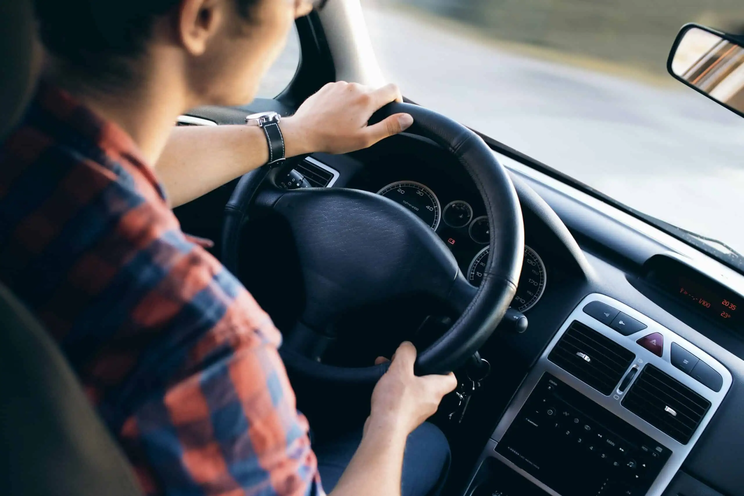 Person driving a car, hands on the steering wheel, dashboard and speedometer visible, wearing a plaid shirt—highlighting the everyday reality for many who face car insurance discrimination or struggle with high rates due to low income.