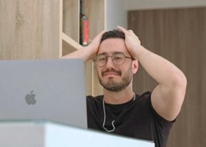 A man wearing glasses holds his head in frustration while looking at a laptop on a desk in a modern room, possibly overwhelmed by concerns about at-will employment Pennsylvania and how employment termination laws PA permit an employer to terminate without reason.