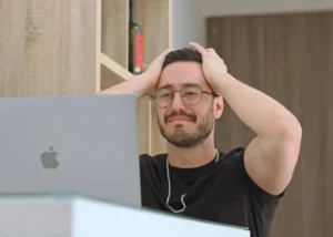 A man wearing glasses sits in front of a laptop, holding his head with both hands and displaying a frustrated facial expression, as he wonders, “Can my employer terminate me without giving a reason?”.