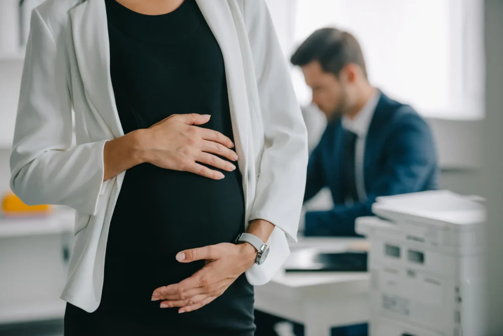 A pregnant woman in business attire stands in an office, holding her belly, as a man in a suit works at a desk—highlighting the environment where a Pregnancy Discrimination Lawyer might offer support.