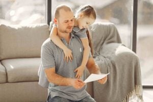 A man sitting on the floor reads a piece of paper—perhaps related to FMLA Leave—while a young girl in a dress hugs him from behind. A couch and blanket are visible in the background.