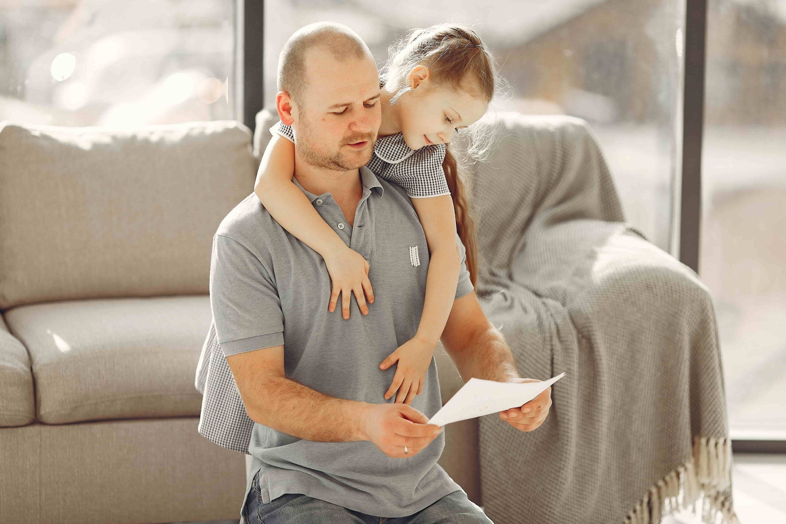 A man sitting on the floor reads a piece of paper—perhaps related to FMLA Leave—while a young girl in a dress hugs him from behind. A couch and blanket are visible in the background.