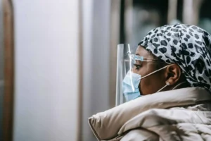 Person in a mask, face shield, and patterned head covering looks ahead indoors, wearing a light-colored coat—perhaps reflecting on questions like Fired On Your Day Off: Can This Really Happen?.