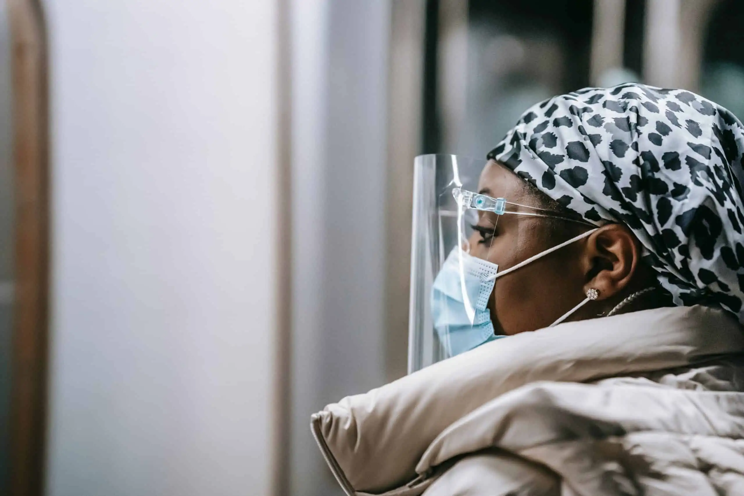 Person in a mask, face shield, and patterned head covering looks ahead indoors, wearing a light-colored coat—perhaps reflecting on questions like Fired On Your Day Off: Can This Really Happen?.