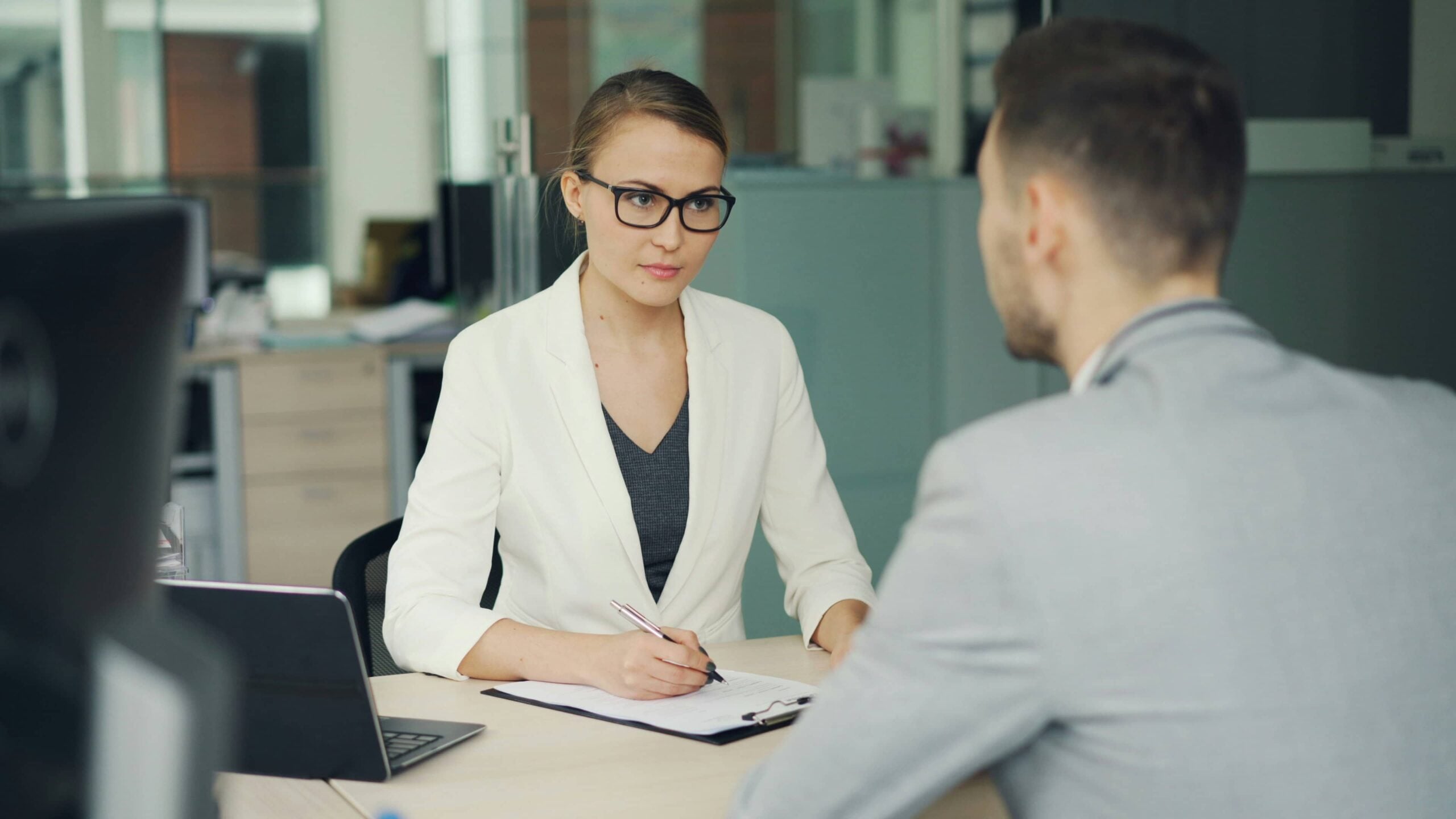 A woman in a white blazer writes on a clipboard while speaking with a man in an office setting, possibly discussing how to file an EEOC complaint New Jersey or whether they should consult a lawyer about their situation.