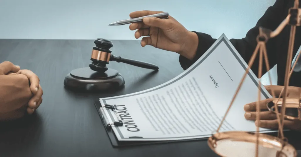 Two people sit at a desk with a gavel, scales, and a clipboard holding a contract; one person gestures with a pen while discussing the document, possibly reviewing details of a Wrongful Termination Case in Philadelphia.