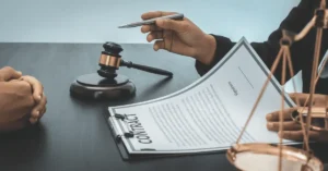 Two people sit at a desk with a gavel, scales, and a clipboard holding a contract; one person gestures with a pen while discussing the document, possibly reviewing details of a Wrongful Termination Case in Philadelphia.