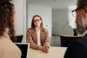 A woman in glasses sits at a table during a meeting or interview with two others in a modern office, possibly discussing topics like "How Much Does a Discrimination Lawyer Cost in New Jersey.