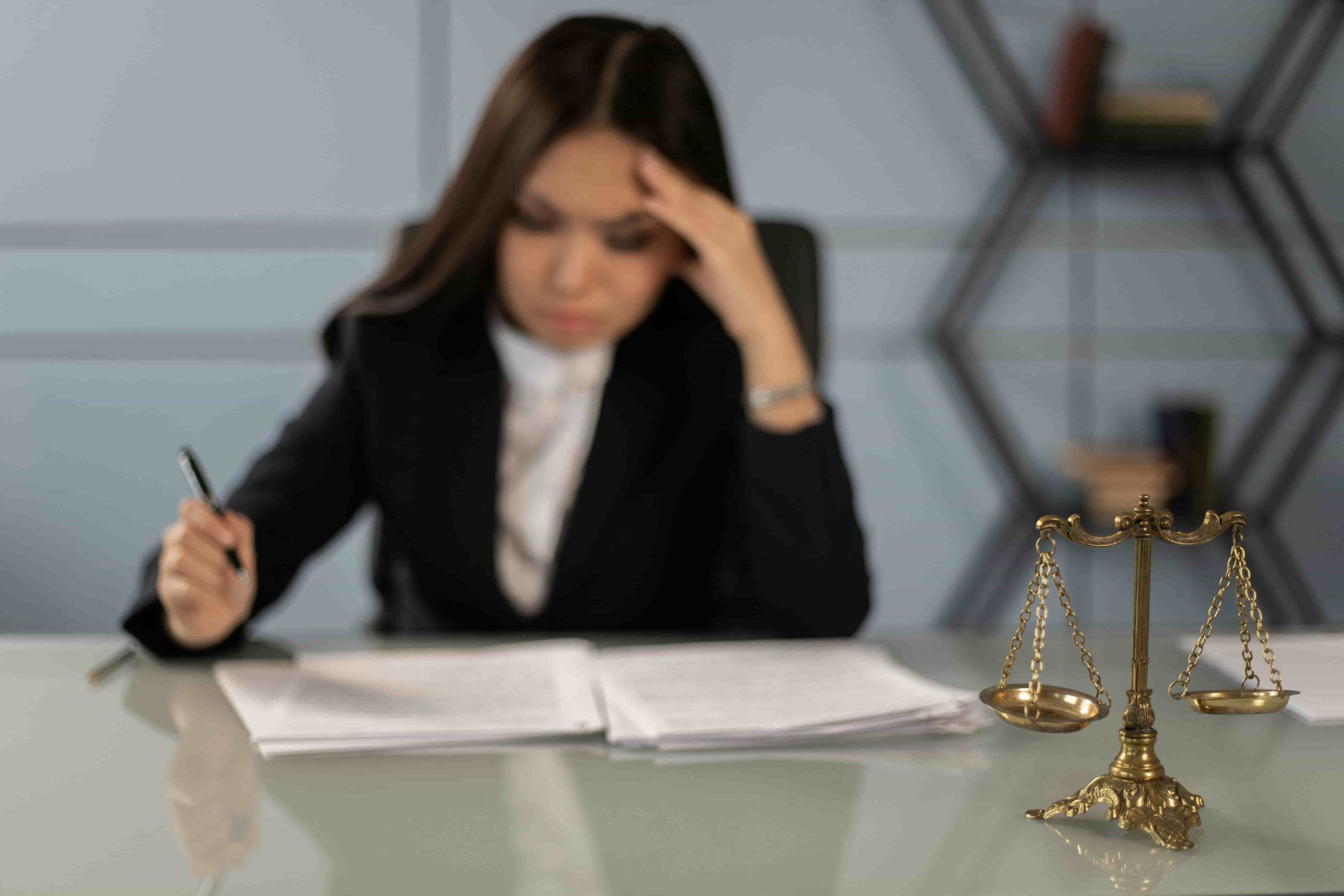 A woman in business attire sits at a desk with legal documents, pondering how to find a top employment discrimination lawyer in New York; a brass scale of justice is featured in the foreground.