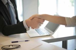 Two people shake hands across a desk with a laptop, a pen, glasses, and papers visible, suggesting a business or professional meeting—such as when consulting an Employment lawyer New Jersey workers trust for workplace guidance.