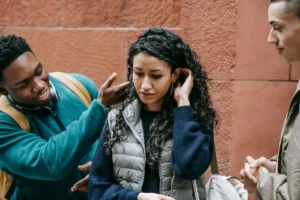 Three people stand together in front of a reddish wall outside; one person looks uncomfortable while another gestures toward them, possibly discussing a serious matter like a sexual harassment case, and the third person stands nearby. The scene suggests the importance of seeking advice—perhaps to hire a lawyer—in situations like this, especially in New Jersey.