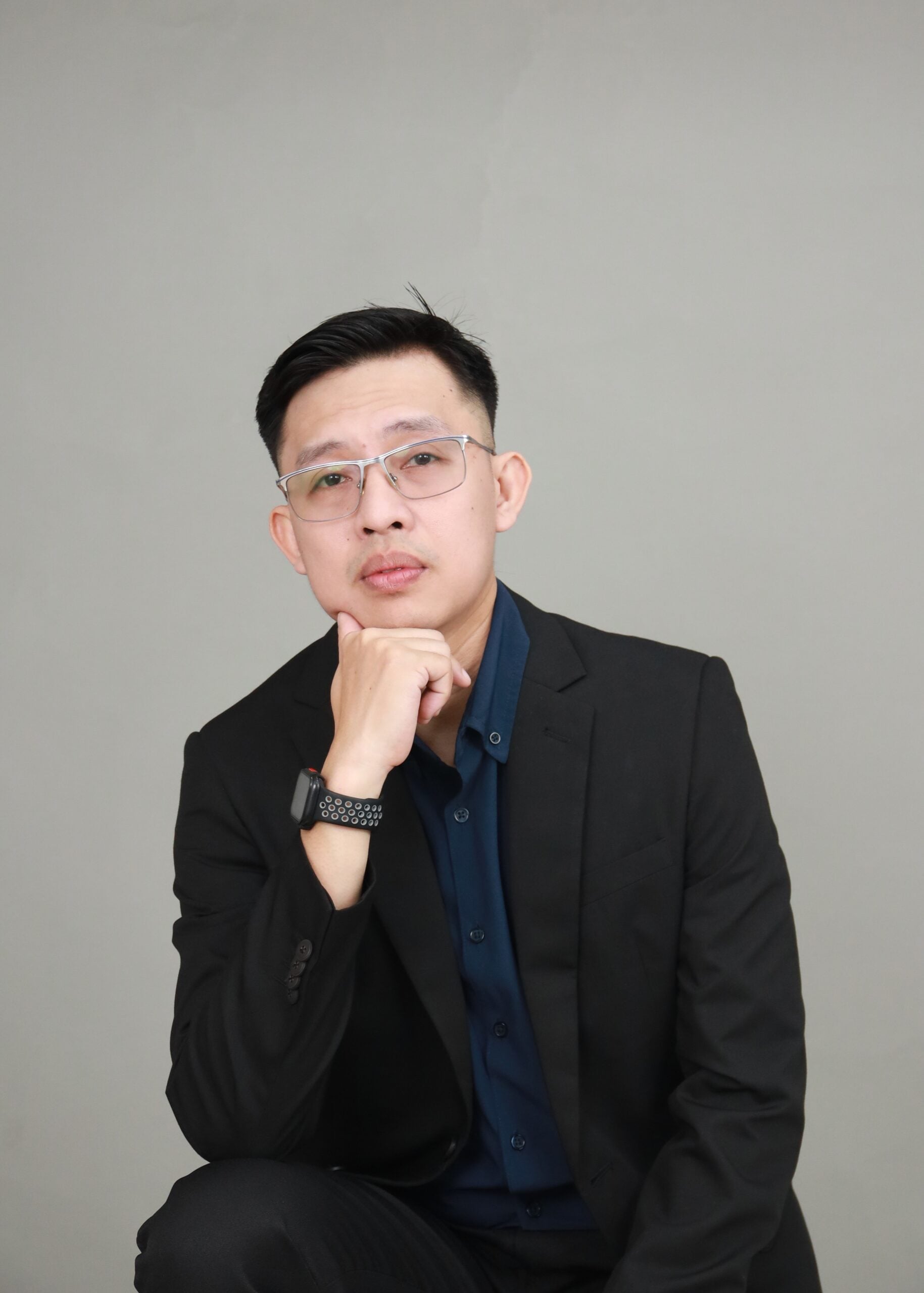Man in a black suit with glasses sits against a plain gray background, resting his chin on his hand and looking at the camera.