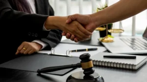 Two people in business attire shake hands across a desk with a gavel, legal documents, a laptop, and scales of justice visible—symbolizing the expertise of Philadelphia Retaliation Lawyers in resolving workplace disputes.