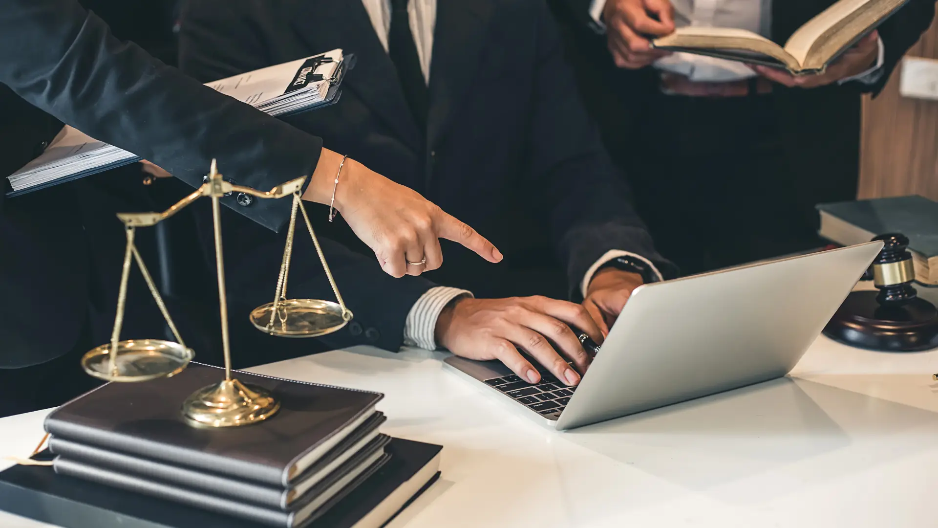 Three people in business attire work at a desk with a laptop, legal books, scales of justice, and a gavel; one person points at the laptop screen, suggesting they may be consulting Philadelphia race discrimination lawyers.