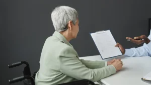 An older woman in a wheelchair sits at a desk across from a person holding a clipboard with documents, highlighting concerns about age discrimination in New Jersey, against a dark background.