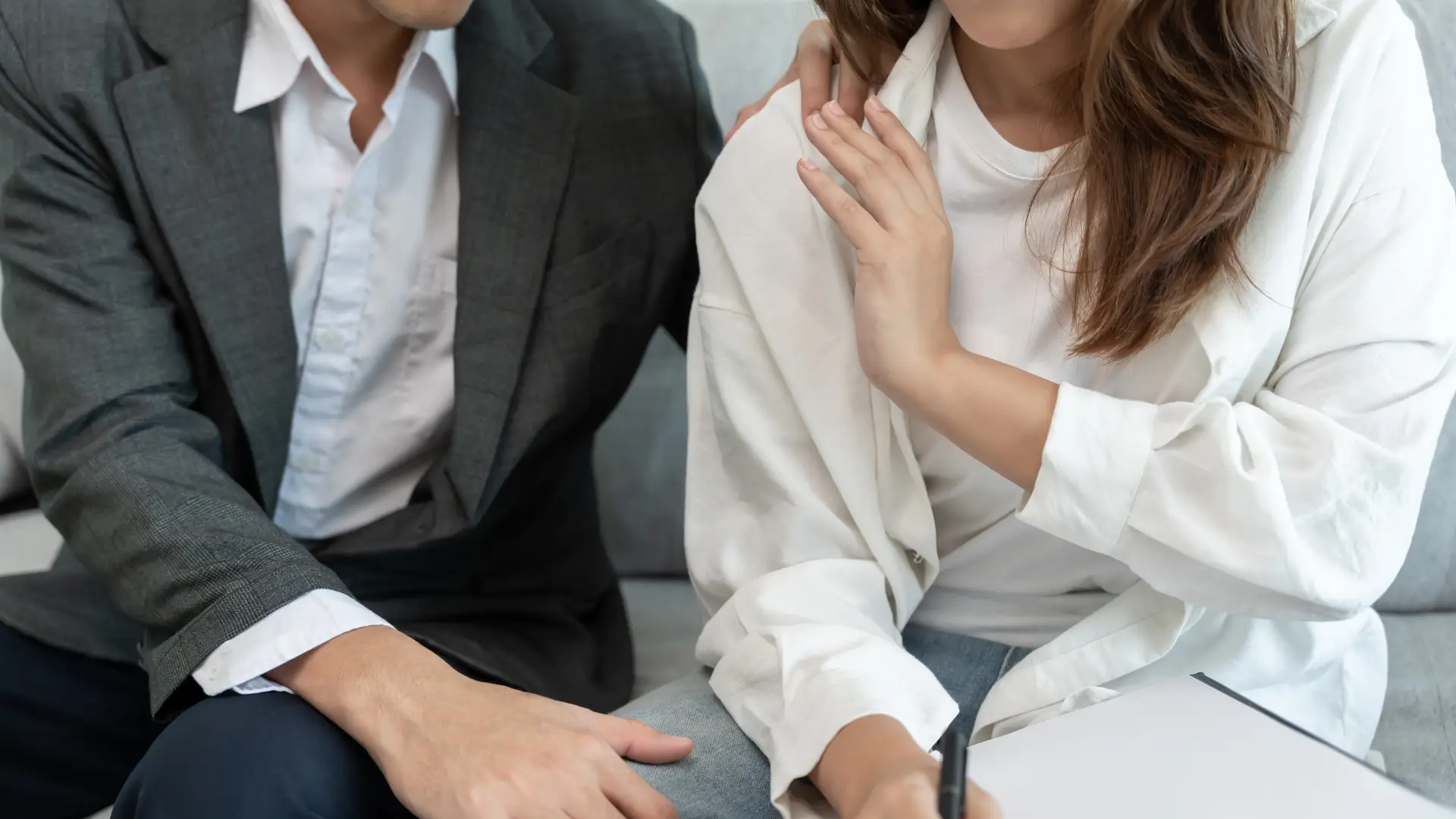 A woman sits beside a man in business attire, touching his shoulder for support as she writes on a clipboard—an image that reflects the compassion and dedication of Philadelphia Sexual Harassment Lawyers.
