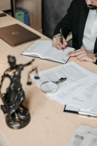A person in business attire writes in a notebook at a desk scattered with legal documents concerning Laws Regarding Unpaid Wages, alongside a laptop, magnifying glass, and a Lady Justice statue.