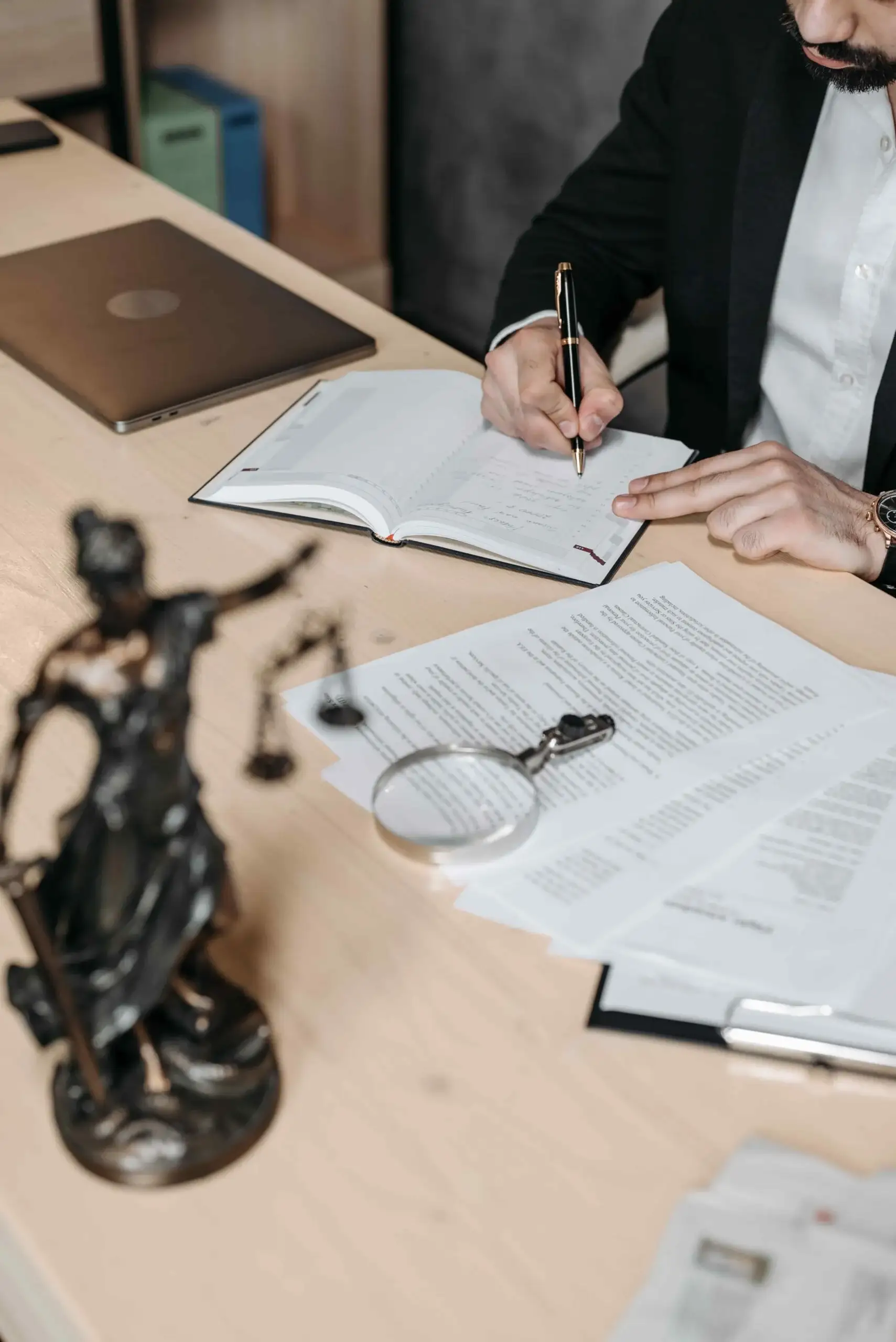 A person in business attire writes in a notebook at a desk scattered with legal documents concerning Laws Regarding Unpaid Wages, alongside a laptop, magnifying glass, and a Lady Justice statue.