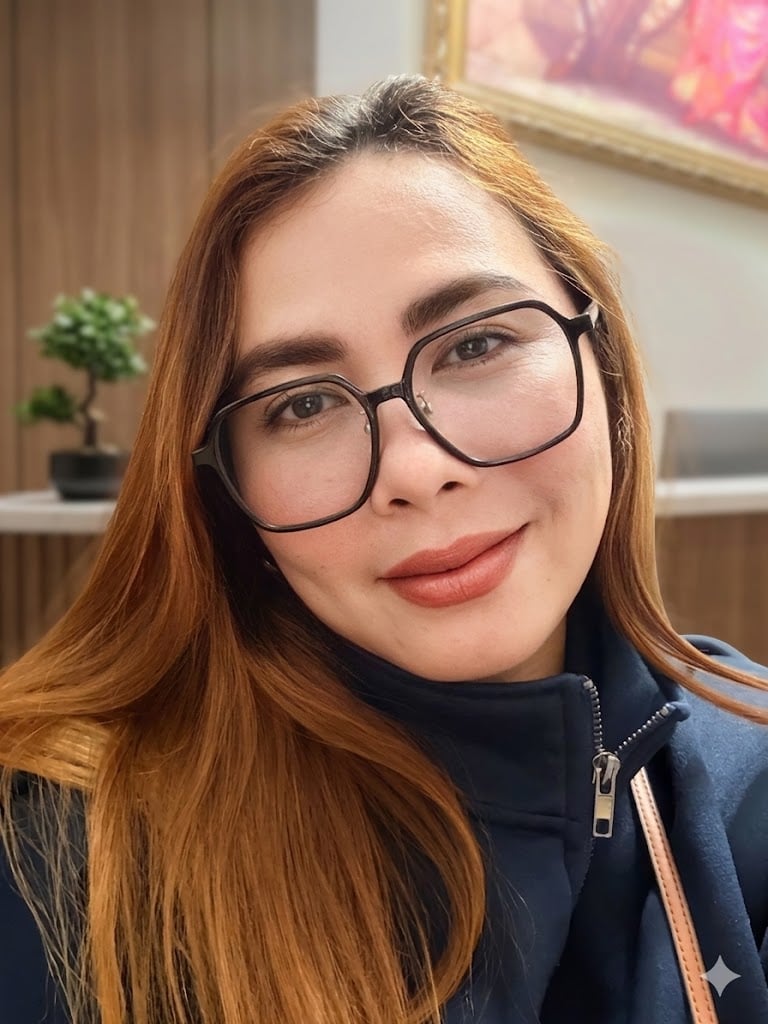 Kat Concepcion, with long light brown hair, wearing glasses and a dark jacket, is looking at the camera indoors with a neutral expression. A small plant and a framed picture can be seen in the background.