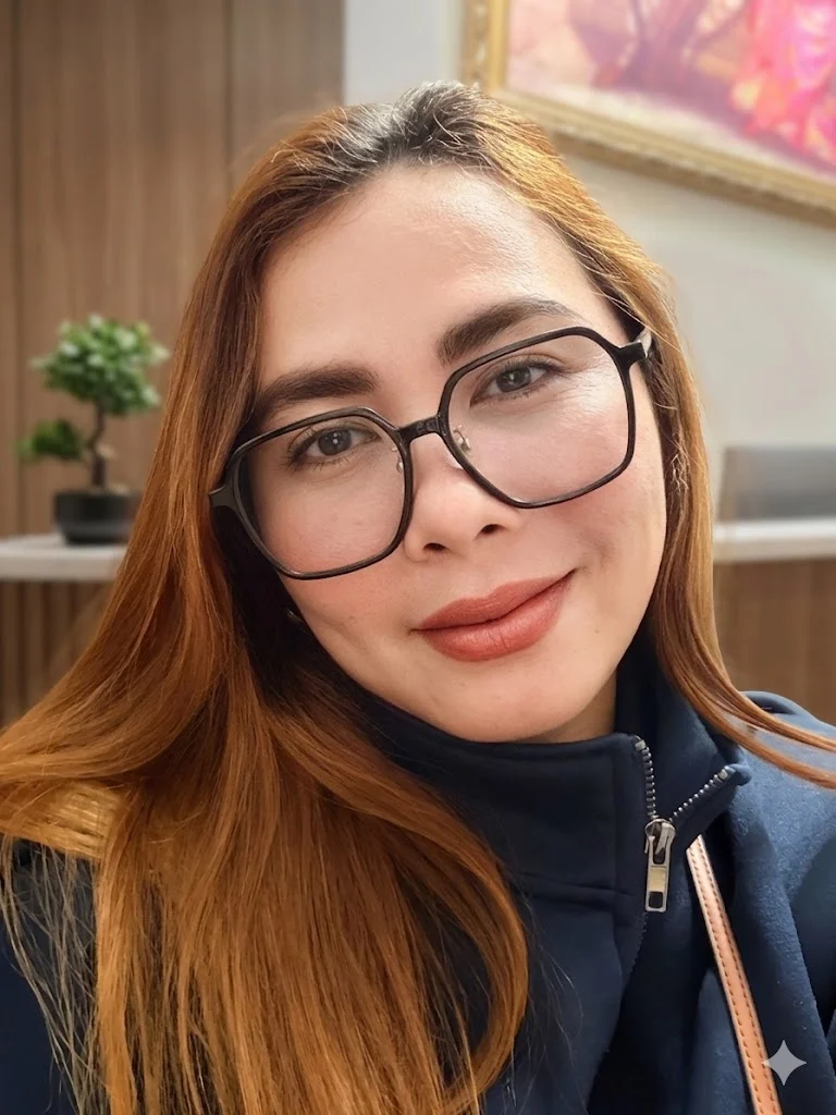 Lorelei Gutilban, a person with long, light brown hair and glasses, smiles at the camera while wearing a dark jacket indoors against a blurred background.