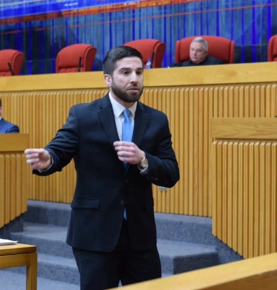 A man in a suit gestures while speaking in a courtroom, representing Lacy Employment Law Firm, with empty red chairs and a judge seated in the background.
