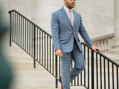 A man in a blue plaid suit with a pink tie walks down marble stairs beside a black railing outside the Lacy Employment Law Firm's white stone building.