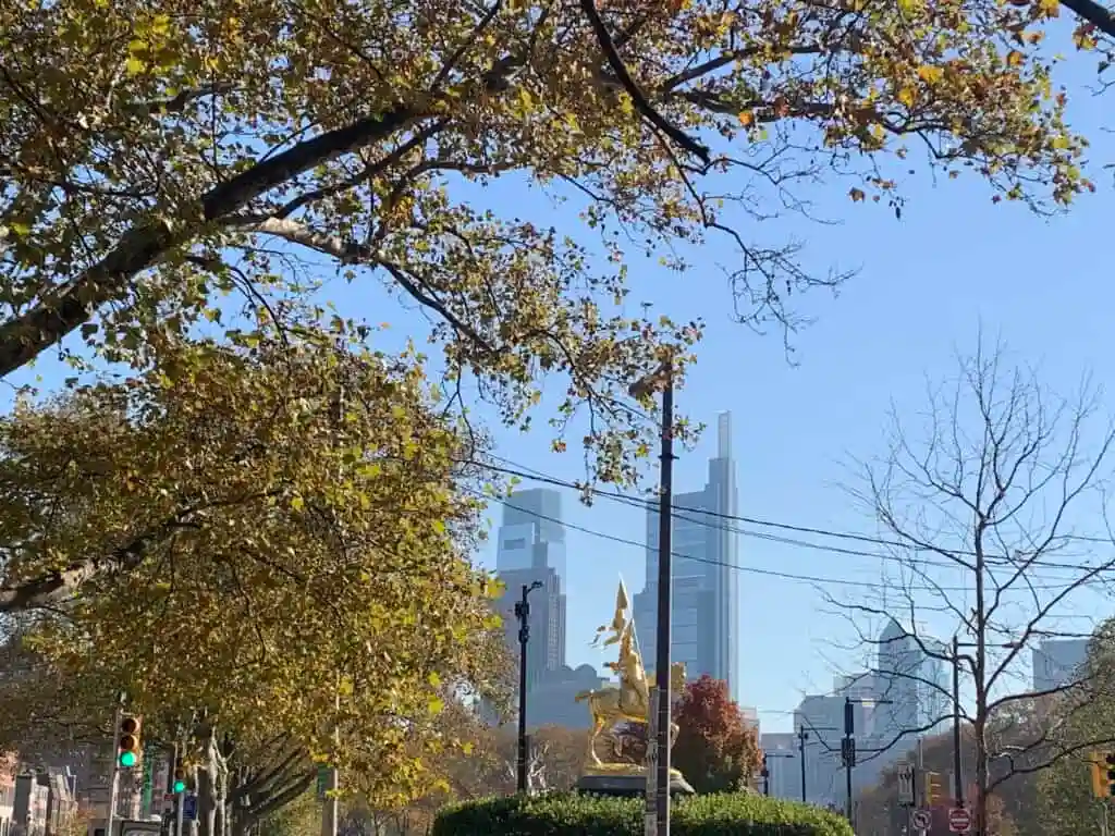 Trees with autumn leaves frame a view of city skyscrapers in the background, a gold statue and traffic lights are visible in the foreground under a clear blue sky—creating an inspiring urban scene that echoes the determination of a dedicated retaliation lawyer standing out amidst the city’s energy and movement.