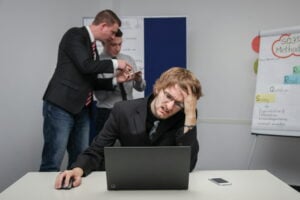 A man sits at a desk in a New Jersey office, looking stressed at his laptop as he tries to collect evidence of workplace discrimination, while two colleagues discuss paperwork in the background.