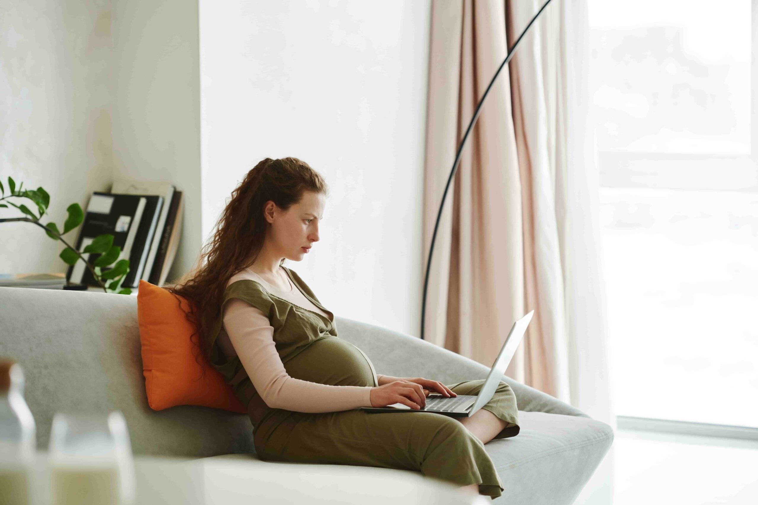 A pregnant woman sits on a couch with an orange pillow, working on a laptop in a bright living room, as she researches pregnant workers rights in New Jersey.