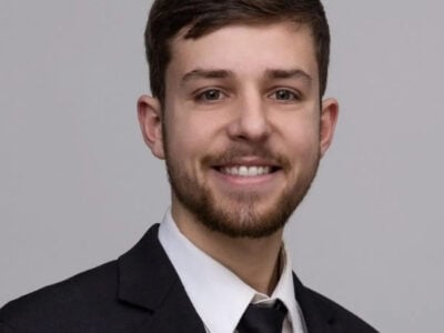 A young man with short brown hair and a beard, wearing a black suit, white shirt, and black tie, smiles at the camera against a plain grey background, representing Lacy Employment Law Firm.