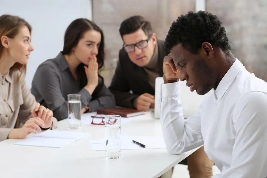 A man sits at a table looking stressed, while three colleagues in the background whisper and look at him. Papers, notebooks, and glasses of water are on the table—a situation where racial discrimination lawyers may be needed.