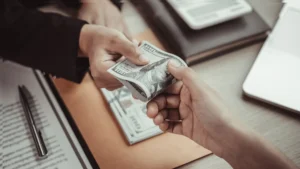 One person hands a $100 bill to another across a desk with documents, a pen, and a laptop—capturing the type of scenario New Jersey Wage and Hour Lawyers often address in workplace legal matters.