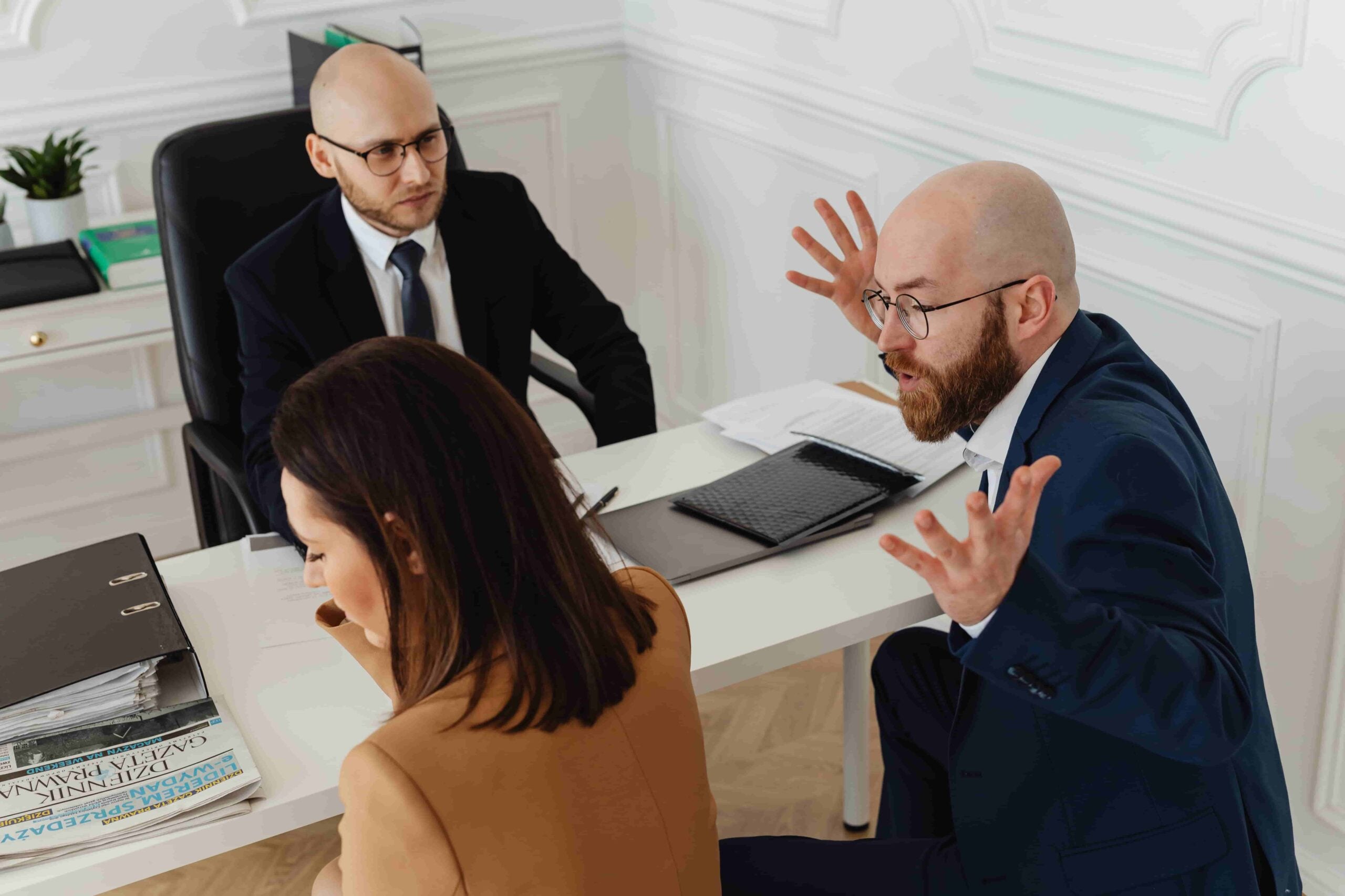 Three people in business attire sit at a desk in an office; one man gestures with raised hands while the others listen, possibly discussing What Is Considered Workplace Retaliation in Pittsburgh?.