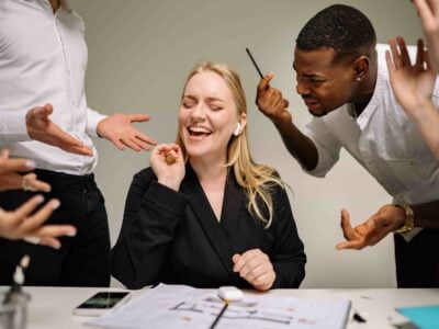 A woman sits at a desk smiling with earbuds in, eyes closed, seemingly tuning out the chaos of a hostile work environment as colleagues around her gesture and speak, appearing frustrated.