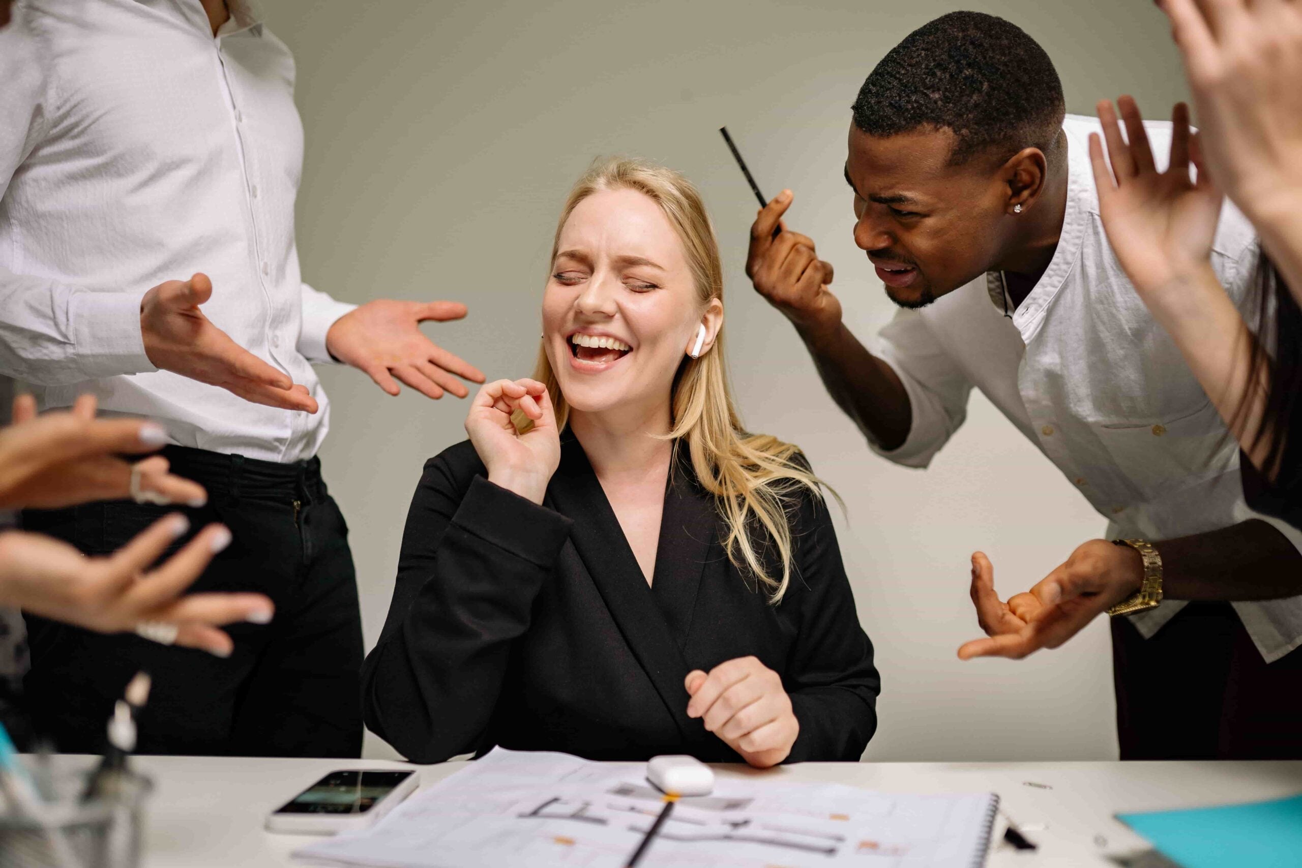 A woman sits at a desk smiling with earbuds in, eyes closed, seemingly tuning out the chaos of a hostile work environment as colleagues around her gesture and speak, appearing frustrated.